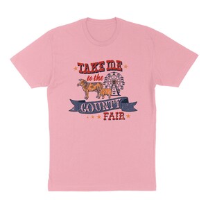 May include: A light pink t-shirt with a graphic design. The design features the text "TAKE ME to the COUNTY FAIR" above a drawing of a cow and a Ferris wheel. The text and images are in red, blue, and brown.
