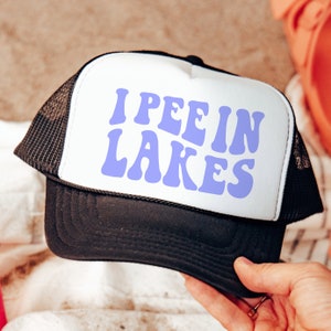 May include: A black and white trucker hat with a white front panel and a black mesh back. The front panel has the text "I PEE IN LAKES" in purple letters.