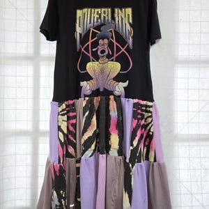 May include: A black t-shirt dress featuring the word "POWERLINE" in a colorful graphic design with a cartoon character. The skirt is a patchwork of various fabrics in shades of purple, brown, and black with tie-dye patterns.