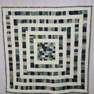 May include: A square quilt with a nested square design. The quilt features a cream-colored background with concentric squares of varying shades of blue, green, and gray fabric squares. The quilt's border is a darker gray.