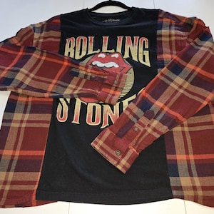 May include: A long-sleeve shirt featuring a black center panel with a distressed Rolling Stones logo and text in yellow and red. The sleeves and side panels are made of a red, tan, and navy blue plaid flannel fabric with buttoned cuffs.