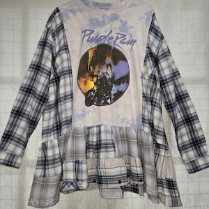 May include: A long-sleeved shirt with a tie-dye top and plaid sleeves and skirt. The shirt features a graphic of a person on a motorcycle with the words "Purple Rain" in a purple font.
