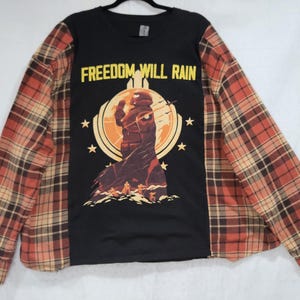 May include: Black long-sleeve shirt with a graphic of a figure in a circle with the text "FREEDOM WILL RAIN" in yellow. The sleeves are a plaid pattern in shades of red, brown, and tan.