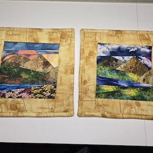 May include: Two square fabric coasters with a landscape design. Each coaster features a central image of mountains, a river, and colorful flowers, set against a blue sky. The borders have a wood-grain pattern.
