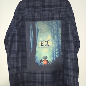 May include: A dark blue plaid flannel shirt with a large graphic on the back featuring the movie title "E.T. The Extra-Terrestrial." The graphic shows E.T. in a forest scene with a glowing heart.