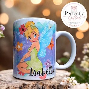 May include: A white ceramic mug featuring a colourful illustration of a blonde fairy with translucent blue wings, wearing a green dress. The name Isabella is printed in black script at the bottom. The background includes blue, red, and orange flowers.