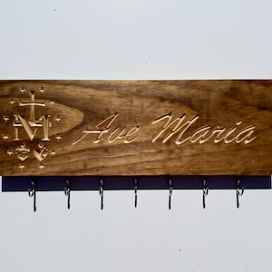 May include: A rectangular brown wooden wall plaque featuring the engraved text Ave Maria in a cursive script. To the left of the text is an engraved symbol consisting of a cross, the letter M, and two hearts surrounded by small stars. Seven metal hooks are attached to the bottom edge.