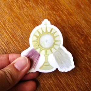 May include: A white sticker with a gold monstrance, a cross, and two white and purple tassels. The monstrance is a religious symbol used in Catholic churches.