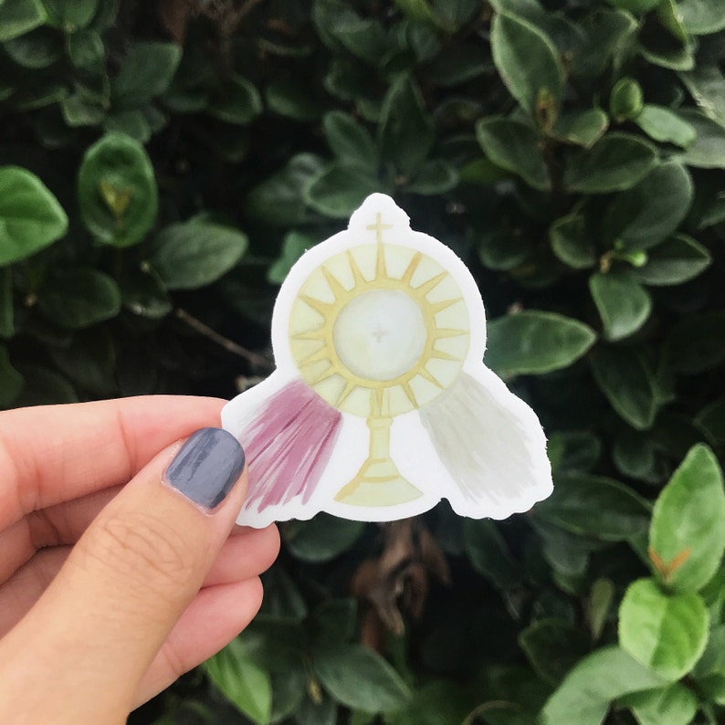 May include: A white sticker with a gold sunburst design, a white cross, and two white and pink tassels. The sunburst has a white circle in the center.