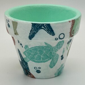 May include: A white terracotta plant pot featuring a hand-painted sea life pattern. The exterior displays teal sea turtles, dark blue starfish, and small bubbles. The interior rim and upper inner wall are painted in a solid, bright mint green color.