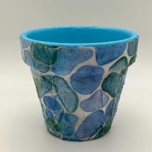 May include: A small, decorative flower pot with a blue interior and a textured exterior. The exterior is decorated with blue and green abstract shapes on a white background. The rim of the pot is painted a bright blue.