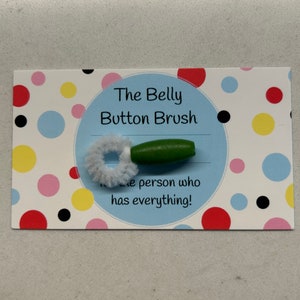 May include: A novelty item, "The Belly Button Brush," is displayed on a polka dot card. The brush features a green wooden handle and a white, fluffy cleaning head. Text on the card reads "The Belly Button Brush" and "...the person who has everything!"
