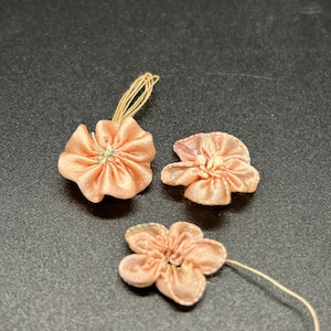 May include: Three small, peach-colored fabric flowers with a gathered center. Two flowers have a thin, beige string attached. The flowers are arranged on a dark surface, showcasing their delicate, petal-like design.