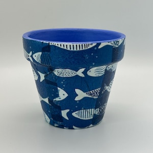 May include: A tapered plant pot featuring a deep blue base color decorated with a repeating pattern of white fish in various styles, including striped and dotted designs. The interior rim and inner walls of the pot are painted a solid bright blue.