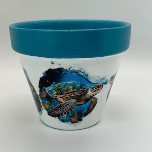 May include: A white plant pot featuring a teal painted rim and a detailed graphic of a sea turtle swimming through blue water with coral reefs. The turtle is depicted in shades of brown, green, and orange, surrounded by small bubbles.