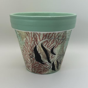 May include: A tapered plant pot with a solid mint green rim and a base featuring a decorative print of black and white angelfish swimming among red coral branches. The design includes subtle map-like text overlays on a light background.