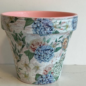May include: A decorative terracotta flower pot with a distressed white background. The pot is adorned with blue hydrangea and pink rose floral patterns. The rim is painted a soft pink. Ideal for indoor or outdoor use.