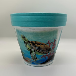 May include: A tapered plant pot featuring a hand-painted scene of a sea turtle swimming near red coral. The upper rim is painted in a solid turquoise color, while the body displays a blue underwater background with a detailed brown turtle.