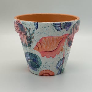 May include: A terracotta plant pot featuring a white exterior decorated with a pattern of colorful seashells, including pink, blue, and purple varieties, along with small blue speckles scattered across the surface of the container.