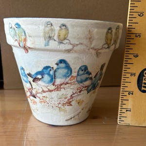May include: A white ceramic flower pot decorated with images of colorful birds perched on branches. The birds are blue, yellow, and orange. The pot is approximately 4 inches tall.