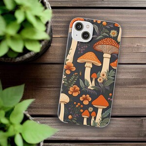 Peut inclure: Une coque de téléphone avec un fond foncé et un motif de champignons et de fleurs orange et blancs. La coque a un découpage pour l'objectif de l'appareil photo blanc.