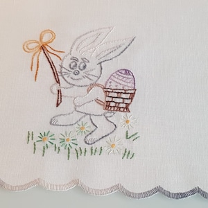 Può includere: Coniglio bianco ricamato che tiene un cestino con un uovo viola e un fiocco giallo. Il coniglio è circondato da erba verde e fiori bianchi.