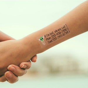 May include: A temporary tattoo applied to a person's arm. The tattoo features a green cartoon character and the text "If I'm lost, please call" followed by phone numbers for Mom and Dad. The tattoo is rectangular and has rounded corners.