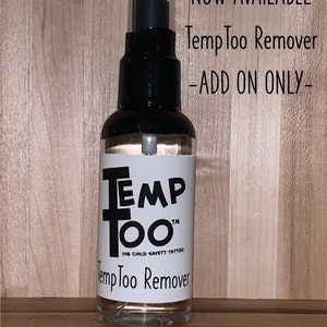 May include: A clear plastic bottle with a black spray nozzle and a white label that reads "Temp Too" and "The Child Safety Tattoo" with the text "Temp Too Remover" below. The bottle is on a wood surface. The text "NOW AVAILABLE Temp Too Remover -ADD ON ONLY- Only available for shipping to the USA." is visible above the bottle.