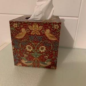 May include: A square tissue box with a decorative pattern featuring birds, flowers, and foliage in shades of red, green, and gold. White tissues protrude from the top. The box is on a glass surface.