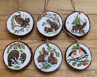 Puede incluir: Conjunto de seis adornos navideños redondos de madera con escenas invernales de animales pintados. Cada adorno presenta un animal diferente: búhos, zorros, ciervos, tejones, conejos y petirrojos. Bordes de madera natural y lazo de cuerda.