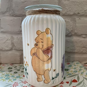 May include: A white, ribbed glass jar with a Winnie the Pooh design. The jar features a cartoon image of Winnie the Pooh, bees, and floral accents. A twine band encircles the top of the jar.
