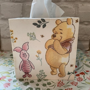 May include: A cream-coloured tissue box decorated with illustrations of Winnie the Pooh and Piglet. The box has floral and bee motifs. White tissues are protruding from the top.