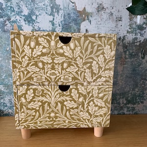 May include: A small wooden chest of drawers featuring two drawers with a mustard yellow and white botanical pattern of oak leaves and acorns. The unit is supported by four short, light-coloured cylindrical wooden legs and has semi-circular cutouts.
