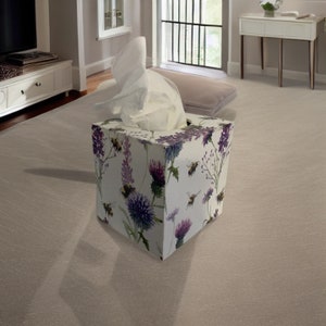 May include: A decorative tissue box with a floral and bee pattern. The box is adorned with purple thistles, lavender, and bees against a white backdrop. White tissues are visible at the top. The box rests on a light-coloured carpet.