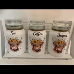 May include: Three white glass jars with lids, labelled "Tea", "Coffee", and "Sugar". Each jar features a Highland cow illustration with a sunflower crown. The jars are displayed on a white shelf.