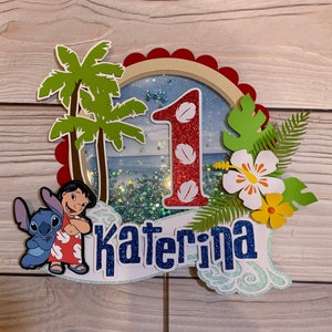 May include: A colorful cake topper featuring a tropical theme with the characters Lilo and Stitch. The number 1 is in red glitter, and the name "Katerina" is in blue glitter. Palm trees, flowers, and a beach scene complete the design.