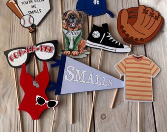 Può includere: Una collezione di oggetti di scena per cabine fotografiche a tema baseball, tra cui una palla da baseball, una mazza da baseball, un guanto da baseball, un berretto da baseball e un gagliardetto con la scritta "SMALLS". Altri oggetti di scena includono un costume da bagno rosso, occhiali da sole e una sneaker nera.