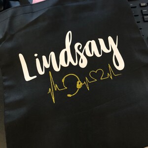 May include: Black tote bag with white cursive text that reads "Lindsay". A gold glitter design of a heartbeat line with a stethoscope and heart is below the name. The bag has black handles.