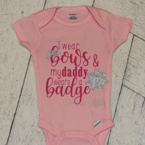 May include: A light pink baby onesie with the text "I wear bows & my daddy wears a badge." The text and decorative bows are in a darker pink color. The onesie is made of soft material.