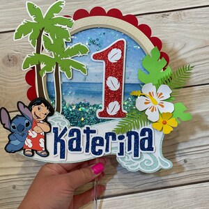 May include: A colorful cake topper featuring a tropical theme with the number "1" and the name "Katerina". The design includes palm trees, a beach scene, and cartoon characters. The topper is made of layered paper and glitter.