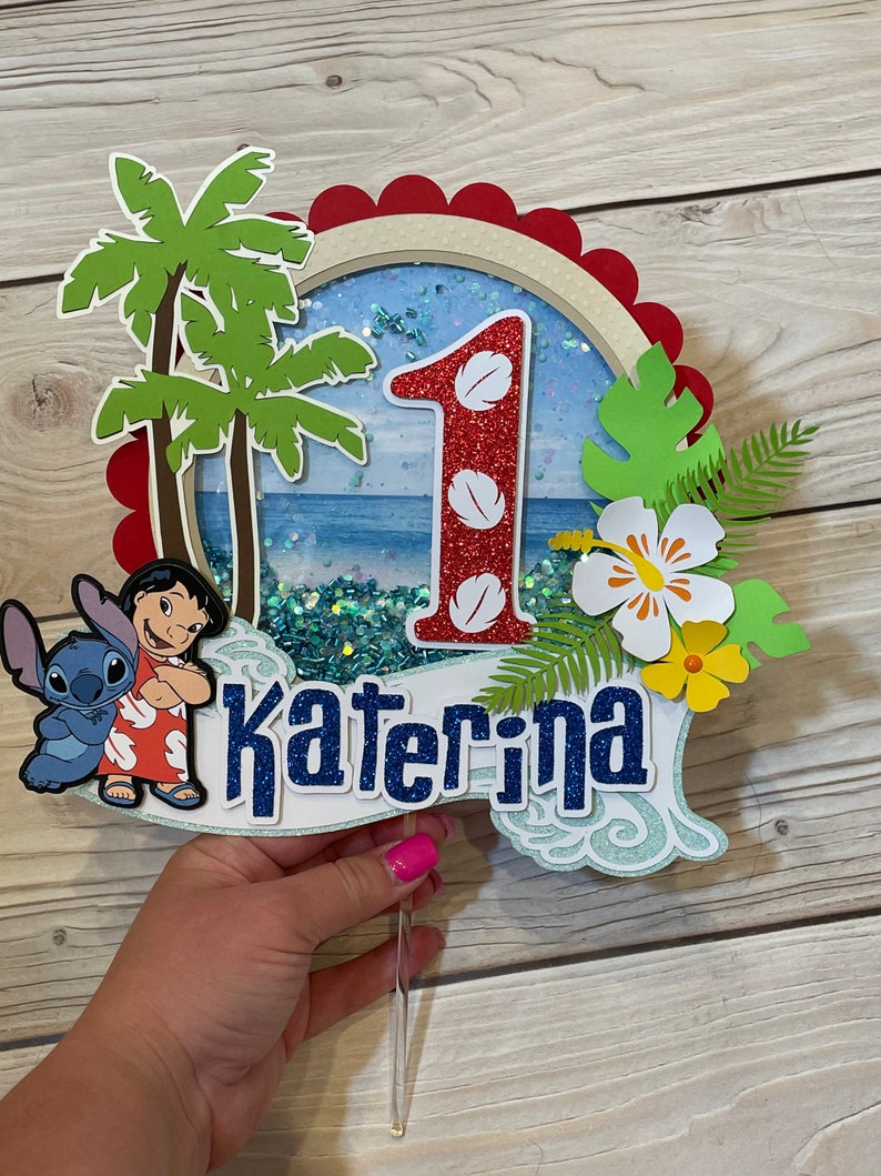 May include: A colorful cake topper featuring a tropical theme with the number "1" and the name "Katerina". The design includes palm trees, a beach scene, and cartoon characters. The topper is made of layered paper and glitter.