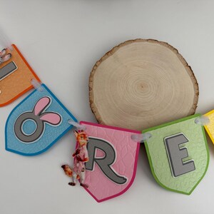 May include: A decorative banner spelling the name LORET with colorful, shield-shaped cardstock pennants. Each pennant features a raised grey letter, textured paper, and cartoon character accents including a rabbit and a fox, all strung together.