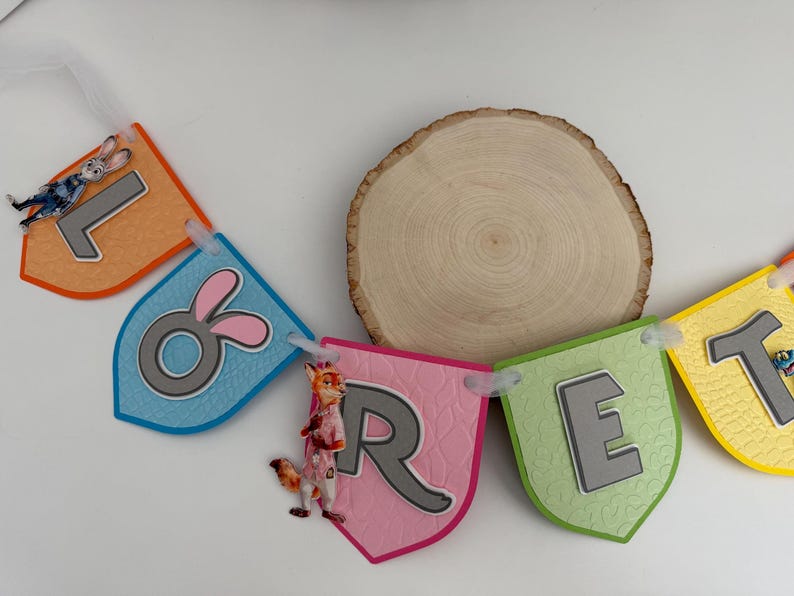 May include: A decorative banner spelling the name LORET with colorful, shield-shaped cardstock pennants. Each pennant features a raised grey letter, textured paper, and cartoon character accents including a rabbit and a fox, all strung together.