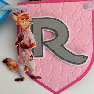 May include: A pink pennant banner piece featuring a textured giraffe-like pattern and a large gray letter R. A cutout of a cartoon fox character wearing a pink floral shirt, a tie, and light-colored pants is attached to the left side of the letter.