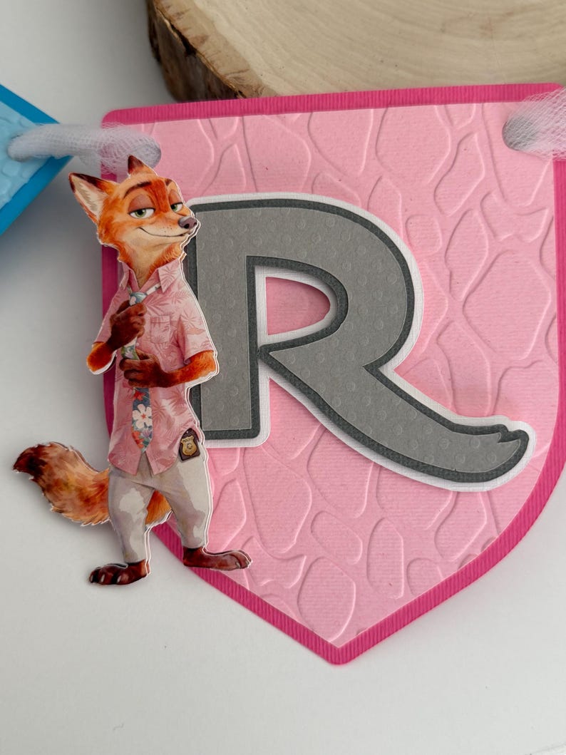 May include: A pink pennant banner piece featuring a textured giraffe-like pattern and a large gray letter R. A cutout of a cartoon fox character wearing a pink floral shirt, a tie, and light-colored pants is attached to the left side of the letter.
