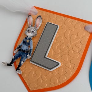 May include: A decorative pennant featuring a peach-colored background with an embossed leopard print pattern, a large gray letter L, and a cutout of a cartoon rabbit character in a blue police uniform, all attached to a white tulle ribbon.