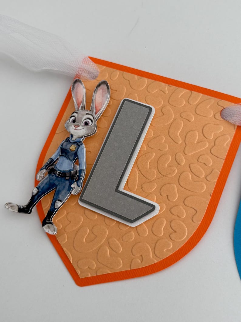 May include: A decorative pennant featuring a peach-colored background with an embossed leopard print pattern, a large gray letter L, and a cutout of a cartoon rabbit character in a blue police uniform, all attached to a white tulle ribbon.
