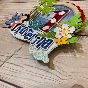 May include: A layered cake topper featuring a tropical theme with the name "Katerina". The design includes a red glitter "1", palm trees, and images of Lilo and Stitch. The topper has a blue and white color scheme with glitter accents.