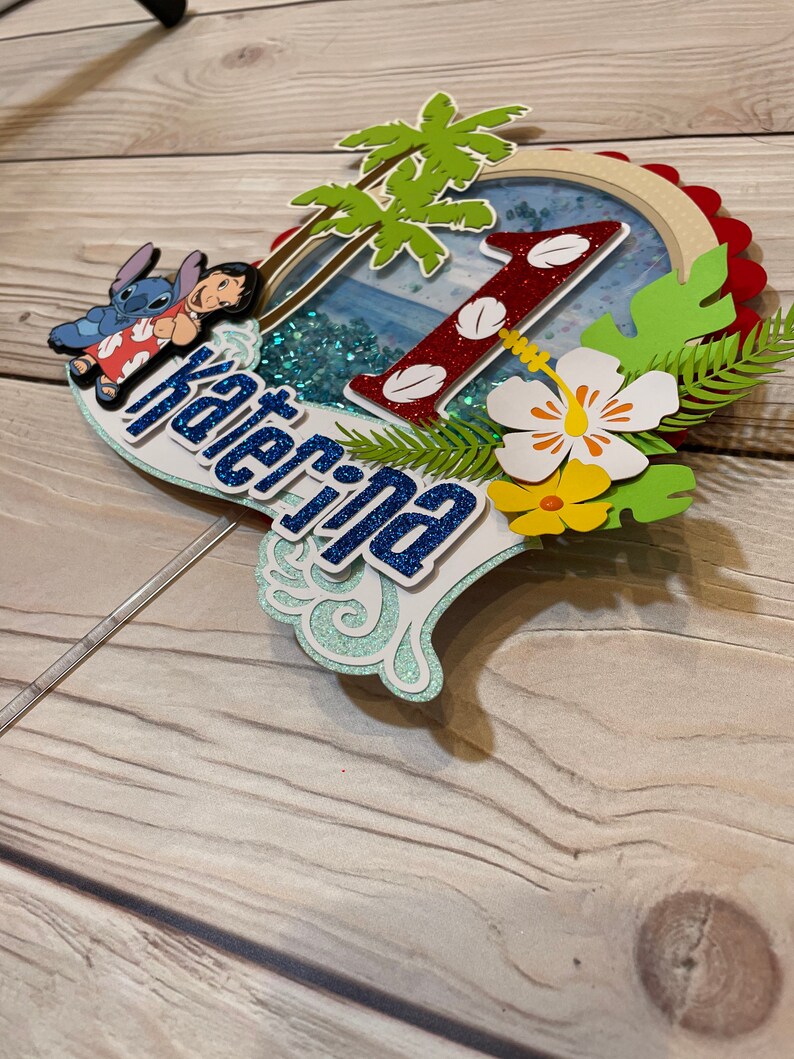 May include: A layered cake topper featuring a tropical theme with the name "Katerina". The design includes a red glitter "1", palm trees, and images of Lilo and Stitch. The topper has a blue and white color scheme with glitter accents.