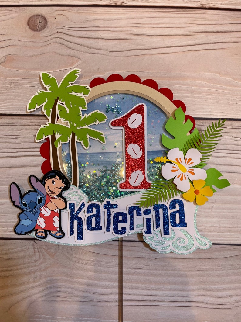 May include: A colorful cake topper featuring a tropical theme with the characters Lilo and Stitch. The number 1 is in red glitter, and the name "Katerina" is in blue glitter. Palm trees, flowers, and a beach scene complete the design.